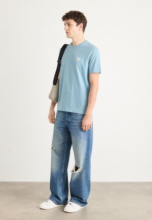 T-shirt bleu clair à manches courtes avec un petit logo, associé à un jean large en denim délavé et des baskets blanches.