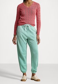 Červený svetr s pleteným vzorem a výstřihem do V, spárovaný se světle zelenými flísovými tepláky s elastickým pasem a manžetami na kotnících, obutý s béžovými teniskami.