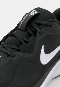 Czarna sportowa buta z fakturowaną cholewką, białym logo Nike, materiałowymi sznurowadłami i kontrastującą gumową podeszwą. Posiada otwory wentylacyjne oraz znakowanie.