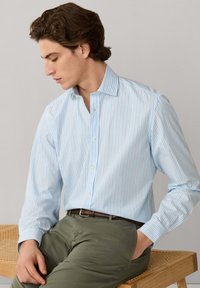 Homme assis sur un banc en bois portant une chemise à rayures bleues claires boutonnée et un pantalon vert olive, regardant vers le bas sur le côté.