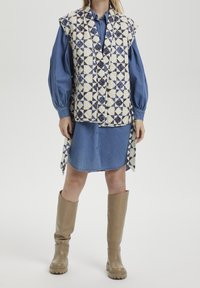Vadderad väst med blå geometrisk mönster över en denimklänning. Knähöga beige stövlar med en slät textur. Övergripande blå och beige färgschema.