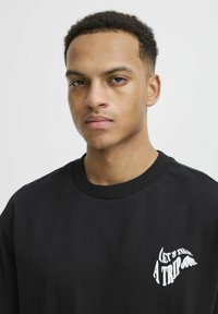 Černý bavlněný sweatshirt s kulatým výstřihem, který má na levé části hrudi bílý text "VYZKOUŠEJME VÝLET". Hladká textura, jednoduchý design.