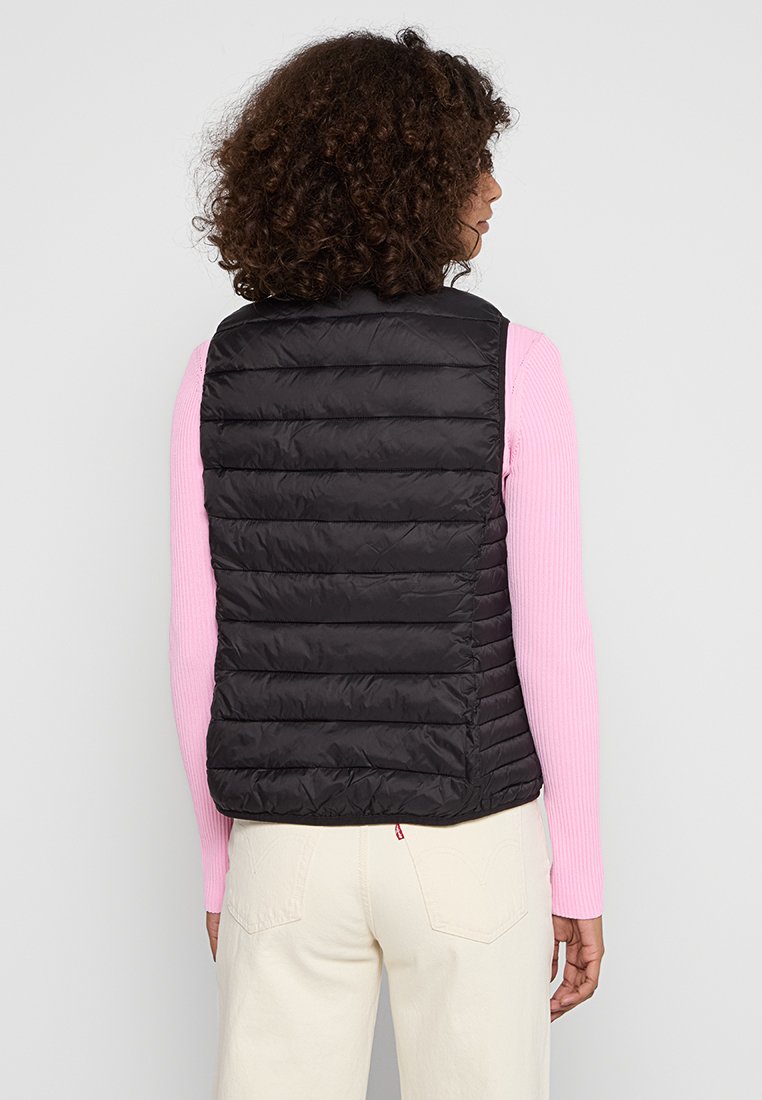 Schwarze Pufferweste mit vertikalen Steppnähten, leichtes Material, ärmelloses Design, getragen über einem pinken Langarmoberteil, einfarbiger Hintergrund.