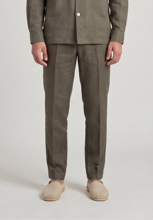 Man draagt olijfgroene op maat gemaakte broek en bijpassend overhemd met beige instapschoenen, staand op een effen achtergrond.