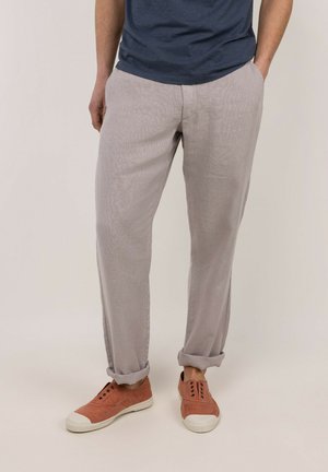 Hombre con pantalones casuales de color gris claro remangados con deportivas sin cordones color óxido y camisa azul oscuro.