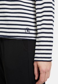 Gestreiftes Hemd aus Baumwolle in Marineblau und Weiß, mit langen Ärmeln und gerippten Bündchen, versehen mit einem kleinen Logo, das nahe dem Saum gestickt ist. Sichtbare schwarze Hose.