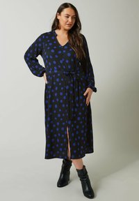 Musta mekko sinisillä pilkuilla, V-pääntie, pitkät hihat, vyötäröltä vyötetty ja edessä halkio. Kangas näyttää olevan teksturoitu. Yhdistetty mustiin nilkkureihin.