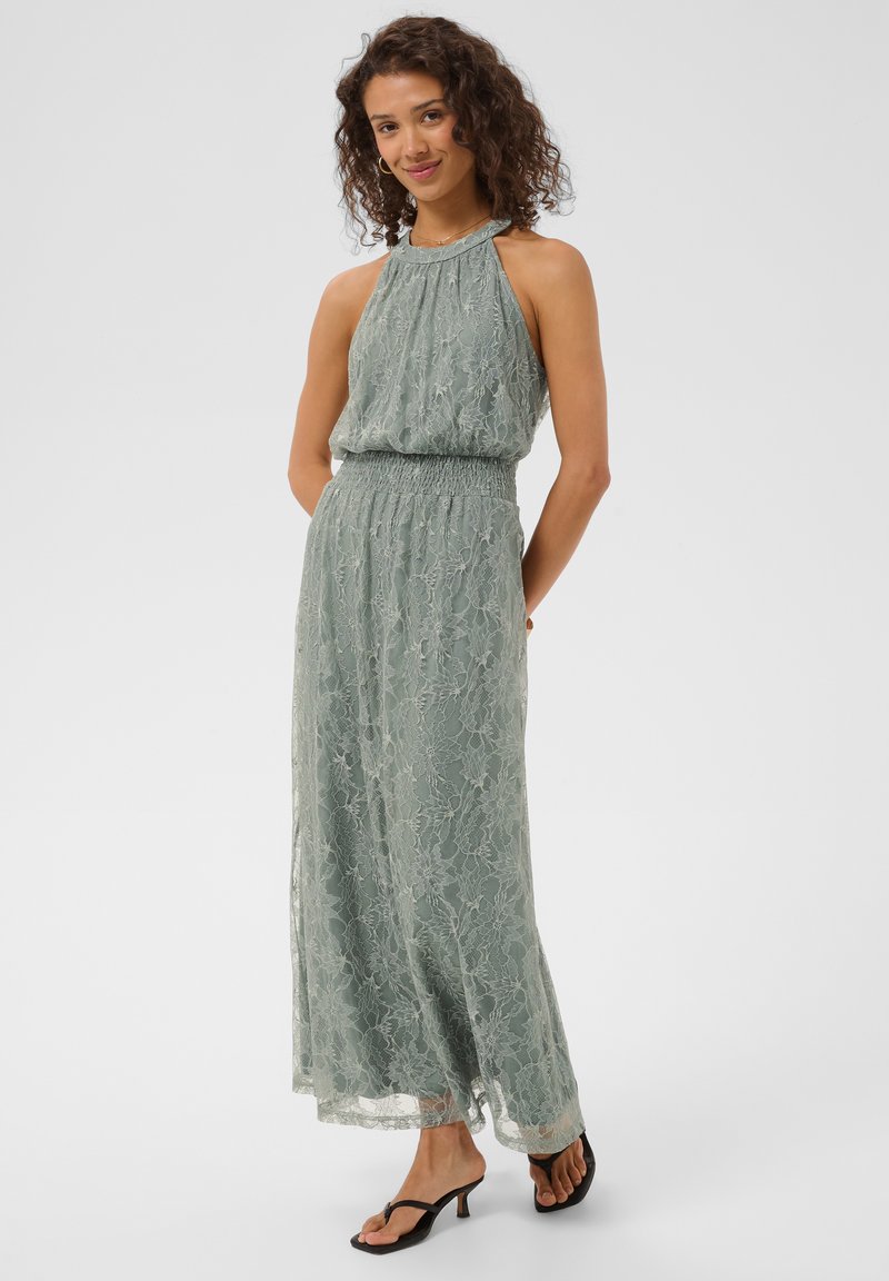 Femme aux cheveux bouclés portant une robe longue en dentelle verte sans manches et des sandales noires à talons, posant les mains dans le dos.