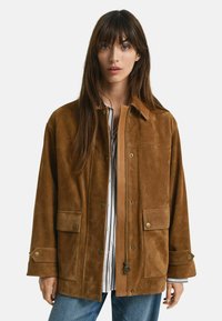 Giacca in suede marrone con due tasche frontali, bottoni a pressione e chiusura con zip. Presenta un colletto e un fit rilassato. Indossata sopra una maglietta a righe.