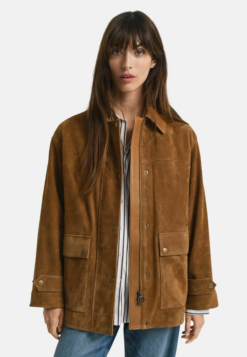 Giacca in suede marrone con due tasche frontali, bottoni a pressione e chiusura con zip. Presenta un colletto e un fit rilassato. Indossata sopra una maglietta a righe.