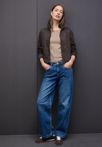 Femme s'appuyant contre un mur gris foncé, portant une veste marron, une chemise beige, un jean large bleu et des baskets marron avec des lacets blancs.