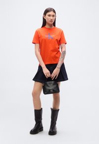 Πορτοκαλί βαμβακερό μπλουζάκι με μπλε λογότυπο Calvin Klein, συνδυασμένο με μαύρη τζιν φούστα και μαύρες μπότες μέχρι το γόνατο, κρατώντας μια μικρή μαύρη τσάντα.