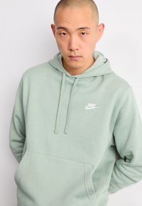 Sweatshirt Nike verde-claro com bolso frontal, capuz ajustável e logótipo branco no peito. Tecido suave e texturizado.