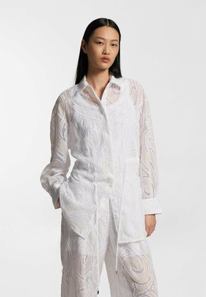 Mujer con cabello largo y negro que lleva un conjunto de camisa y pantalones blancos translúcidos con patrones bordados en espiral, de pie contra un fondo claro y sencillo.