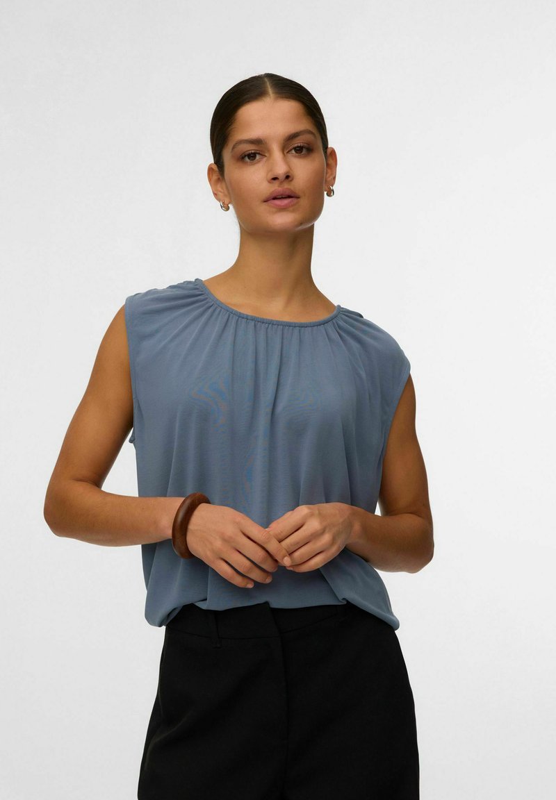 Femme aux cheveux foncés attachés en chignon, portant un chemisier bleu sans manches et un pantalon noir, debout avec les mains légèrement jointes, sur un fond uni.