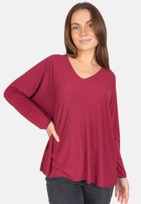 Haut à manches longues bordeaux en V, en tissu doux, avec épaules tombantes et fentes latérales pour plus de liberté de mouvement, associé à un jean foncé.