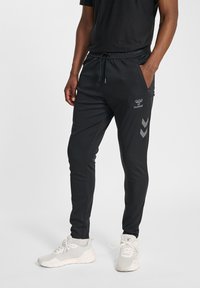 Schwarze, sportliche Jogginghosen mit Kordelzugbund, Seitentaschen und grauem Logo. Seitlich mit eckigen grauen Chevron-Mustern verziert.