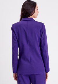 Blazer violet avec une coupe ajustée, des revers nets et une texture lisse. Dispose d'épaules structurées et d'une fente arrière unique pour une plus grande liberté de mouvement.