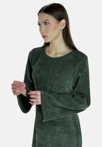 Zielona sukienka z długim rękawem, wykonana z teksturowanej tkaniny, z okrągłym dekoltem i rękawami w kształcie dzwonu, z widocznymi detalami szycia.