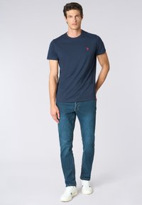 T-shirt in cotone blu navy con maniche corte, colletto rotondo e piccolo logo rosso sul petto. Abbinato a jeans blu e sneakers bianche.