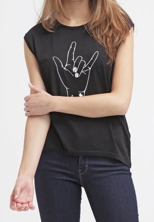 Frau trägt ein schwarzes ärmelloses Shirt mit weißem Motiv einer Hand, die auf Gebärdensprache das Zeichen "Ich liebe dich" macht, kombiniert mit dunklen Jeans.