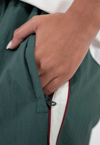 Main de ton de peau moyen insérée dans la poche d'un pantalon vert foncé avec bordure blanche et rouge et tirette de fermeture éclair visible.