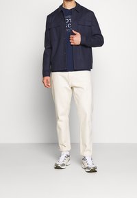 Granatowa koszula z guzikami i dwiema kieszeniami na piersi, noszona na ciemnym T-shircie, w zestawieniu z kremowymi spodniami i wielokolorowymi sneakersami.