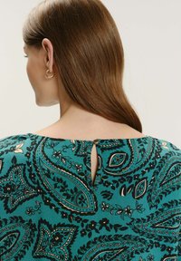 Mujer con cabello castaño liso que lleva pendientes de aro dorados y una blusa de estampado paisley en tonos turquesa con cierre de botones en la parte posterior del escote.