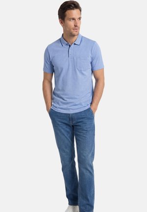 Uomo in piedi con le mani nelle tasche, indossa una polo azzurro chiaro, jeans blu e scarpe bianche, guarda leggermente di lato.