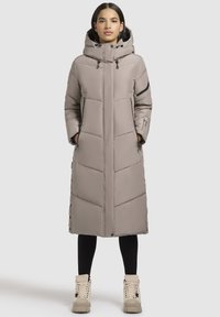 Garš beigžņotā pūkā jaka ar kapuci, ar rāvējslēdzīgu kabatu, quiltota raksta un pogu aizdarēm sānā. Kombinēta ar gaišiem zābakiem.