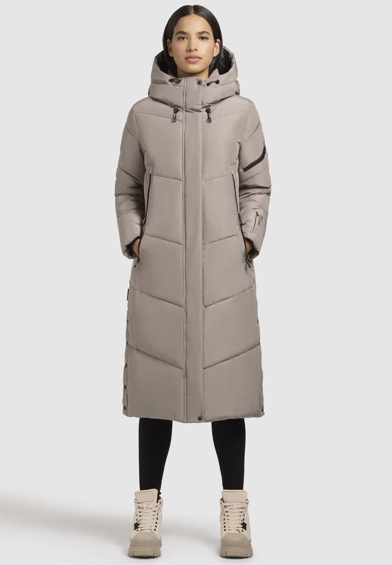 Garš beigžņotā pūkā jaka ar kapuci, ar rāvējslēdzīgu kabatu, quiltota raksta un pogu aizdarēm sānā. Kombinēta ar gaišiem zābakiem.