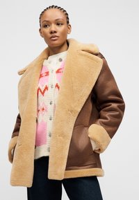 Γυναίκα που φοράει καφέ μπουφάν με εσωτερική επένδυση από γούνα πάνω από λευκό ζακέτα με ροζ και κόκκινα σχέδια, στέκεται με τα χέρια στις τσέπες του μπουφάν.