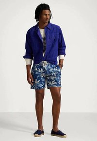 Μπλε καμβάς μπουφάν, λευκό μπλουζάκι με γραφικό σχέδιο, μπλε λουλουδάτα σορτσάκια μπάνιου και ναυτικά εσπαντρίγιες, με ελαστική μέση και σχέδιο με φύλλα φοινίκων.