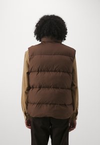 Braune Pufferweste mit hohem Kragen, die gesteppten Abschnitte und eine glatte, glänzende Oberfläche aufweist, getragen über einem beigen Langarmshirt.