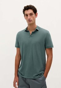 Kortærmet polo shirt i dæmpet grøn, med en blød tekstur, lille krave og lige kant. Parret med grå shorts.