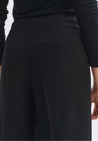 Pantalon texturé noir avec une coupe ajustée, doté d'une poche arrière et d'une ceinture lisse. Le tissu présente un motif diagonal subtil.
