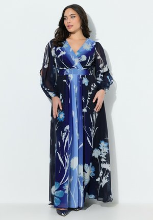 Frau trägt ein langes, fließendes blaues Blumenkleid mit transparenten, aufgeblähten langen Ärmeln, steht vor einem schlichten weißen Hintergrund.