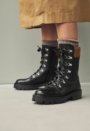 Las botas de cuero negro con cordones presentan herrajes plateados, una suela de goma gruesa y un acento de gamuza marrón en el tobillo. Se pueden ver calcetines de lana texturada.