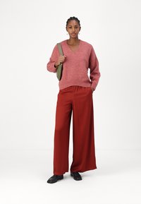 Pullover di lana rosa con scollo a V, abbinato a pantaloni a gamba larga color ruggine. Il modello tiene una piccola borsa verde oliva e indossa scarpe nere.