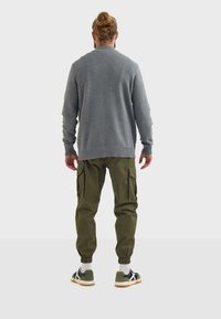 Maglione grigio lavorato a maglia, pantaloni cargo verdi con tasche laterali e sneakers a fantasia, visti da dietro su uno sfondo neutro.