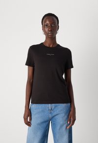 Svart bomull T-skjorte med rund hals, korte ermer og en liten hvit logo-tekst på forsiden. Kombinert med lyseblå denimbukser.