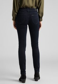 Jeans skinny de color azul oscuro con una textura suave, cintura media y dos bolsillos traseros. Llevados con botas negras de altura al tobillo y una blusa verde.