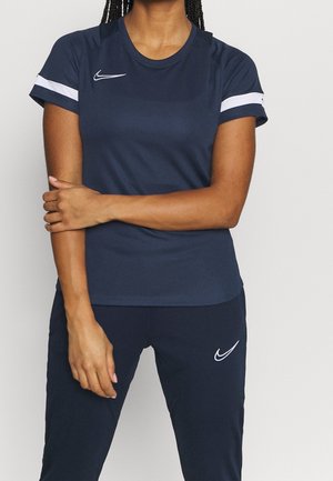 Mujer con el cabello trenzado, vestida con ropa deportiva azul marino de Nike con rayas blancas en las mangas y logotipos en el pecho y los pantalones, de pie con los brazos cruzados.
