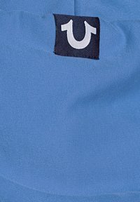 Étiquette de vêtement noire avec un logo blanc en forme de fer à cheval sur un tissu bleu texturé.