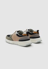 Par de zapatillas beige, verdes, negras y marrones con suelas blancas gruesas, colocadas de espaldas sobre un fondo gris.