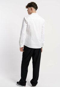 Biała koszula z długim rękawem, z kołnierzykiem, gładka w dotyku, z guzikami przy mankietach, zestawiona z luźnymi czarnymi spodniami i czarnymi butami.