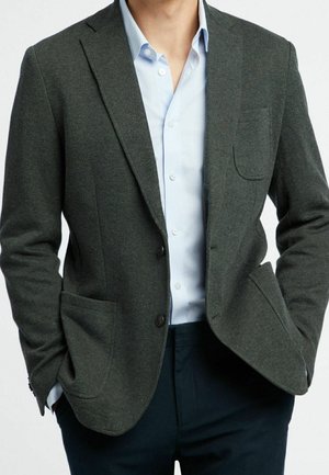 Mann trägt einen dunkelgrauen Blazer über einem hellblauen Hemd mit Knöpfen, die Hände in den Taschen und dunkle Hose.