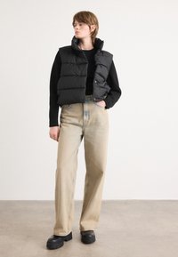 Colete puffer preto com gola alta e botões de pressão, combinado com jeans de perna larga bege claro. Sapatos plataforma pretos completam o visual.