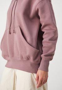 Felpa rosa con tasca frontale, cappuccio con cordino, polsini e orlo a coste. Tessuto morbido, vestibilità leggermente ampia, indossata sopra pantaloni chiari.