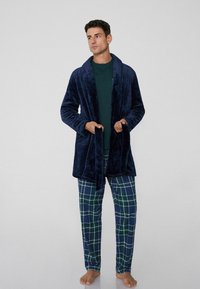 Tmavě modrý plyšový župan s šálovým límcem, zavazováním v pase a postranními kapsami, nošený přes zelený top a kostkované pyžamové kalhoty v zelené a tmavě modré barvě.